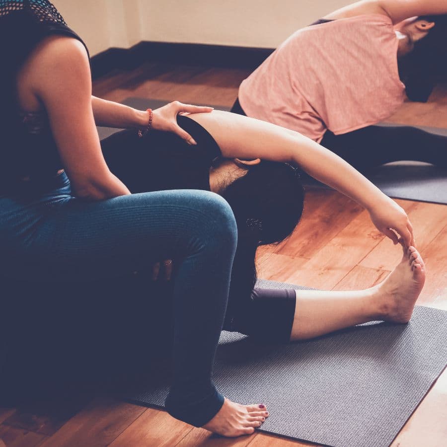 Yoga teacher assisting a student in a seated forward fold